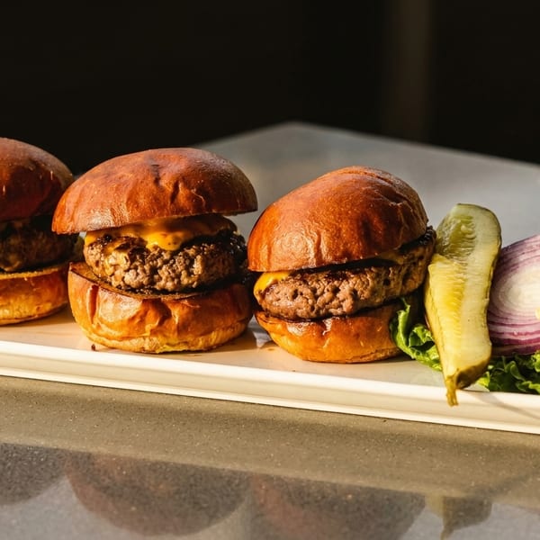 Three cheeseburger sliders on brioche buns with a side of lettuce, tomato, red onion, and a pickle.