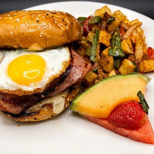 Breakfast sandwich with fried egg, ham, and cheese, served with potatoes, peppers, and fruit.