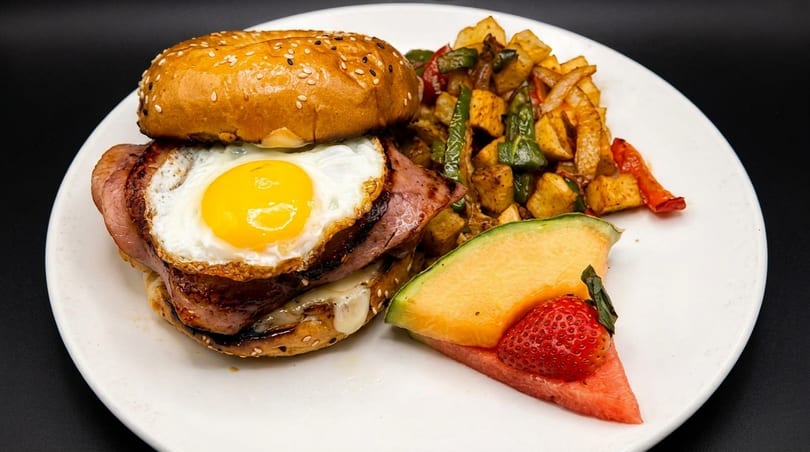 Breakfast sandwich with fried egg, ham, and cheese, served with potatoes, peppers, and fruit.