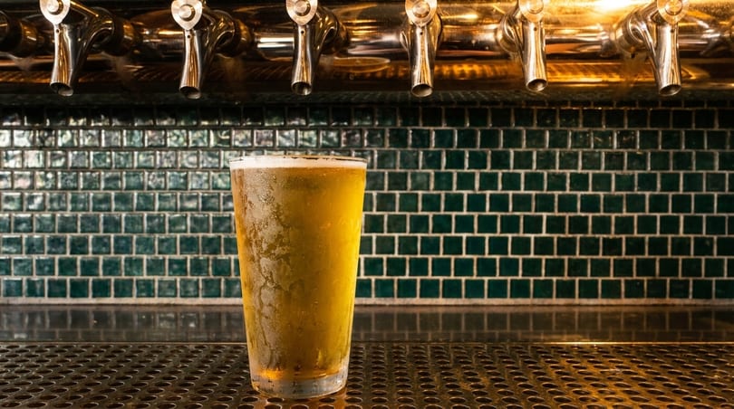 A frosty glass of beer sits on a bar counter with beer taps and green tiled wall in the background.