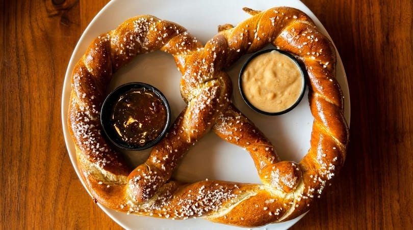 A large, salted pretzel with two dipping sauces, one dark and one light, on a white plate.