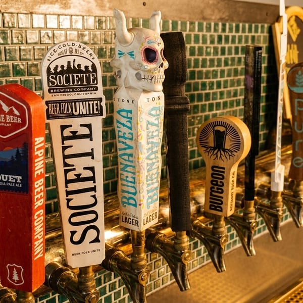 A row of diverse beer tap handles, including Alpine Beer, Societe, and Buenaveza, against a green tiled wall.