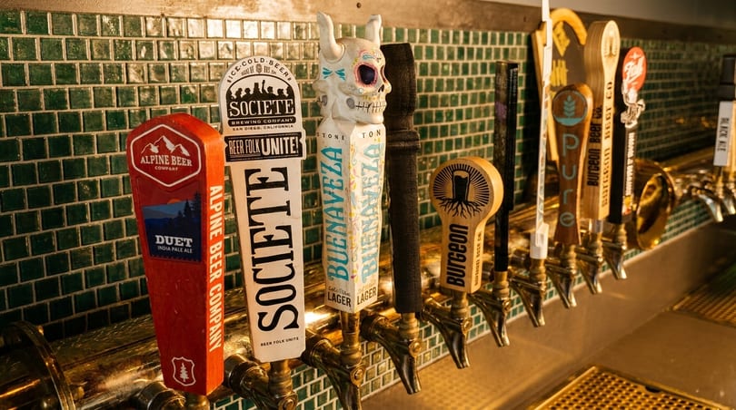 A row of diverse beer tap handles, including Alpine Beer, Societe, and Buenaveza, against a green tiled wall.