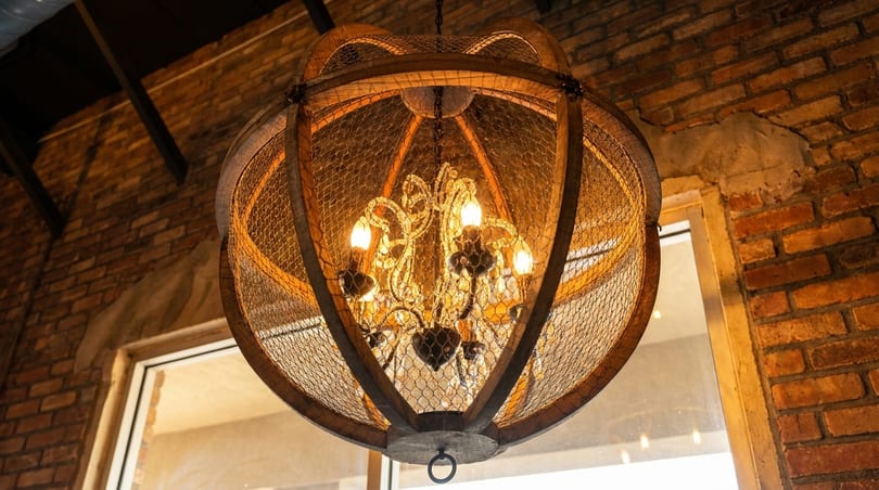 A rustic chandelier with a chicken wire and wood frame hangs in a brick-walled room.
